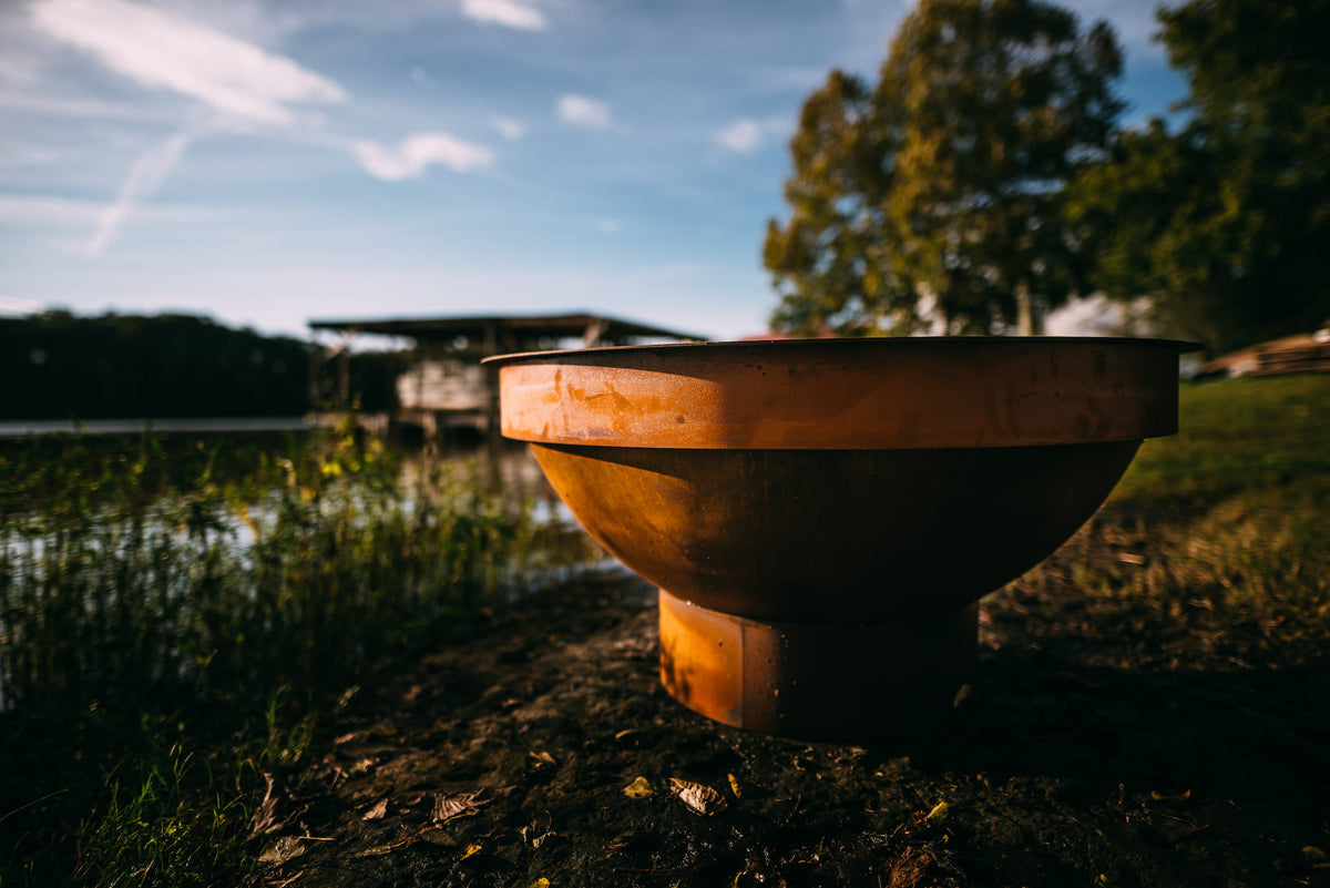 Sideview of fire pit next to lake. The fire pit has a Fire Pit Art 40” Carbon Steel Table Top Lid covering it.