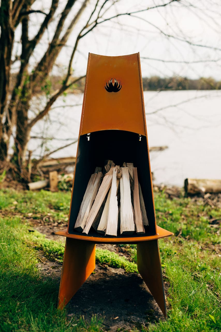 Orange portable stove with wood inside, set against a natural background with trees and water.