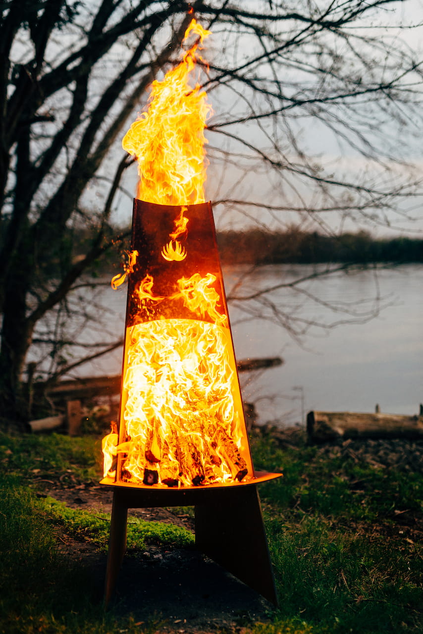 Flaming fire pit sculpture in a natural setting with trees and water in the background.