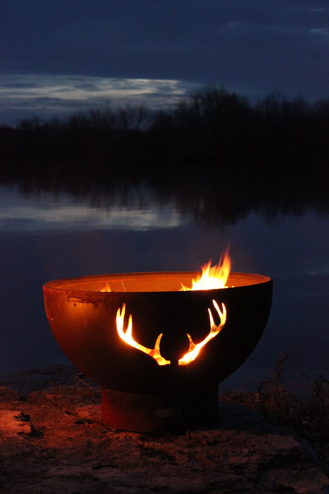 Rustic Antlers 36" Steel Fire Pit by Fire Pit Art glowing at night with serene lake reflection. Available in wood burning, natural gas, or propane.