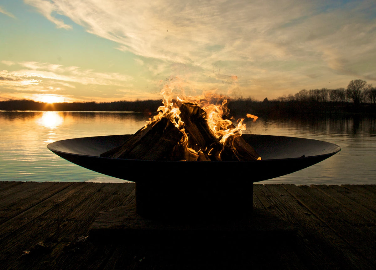 Asia Steel Fire Pit, 72", with flames burning on a dock during sunset over water.