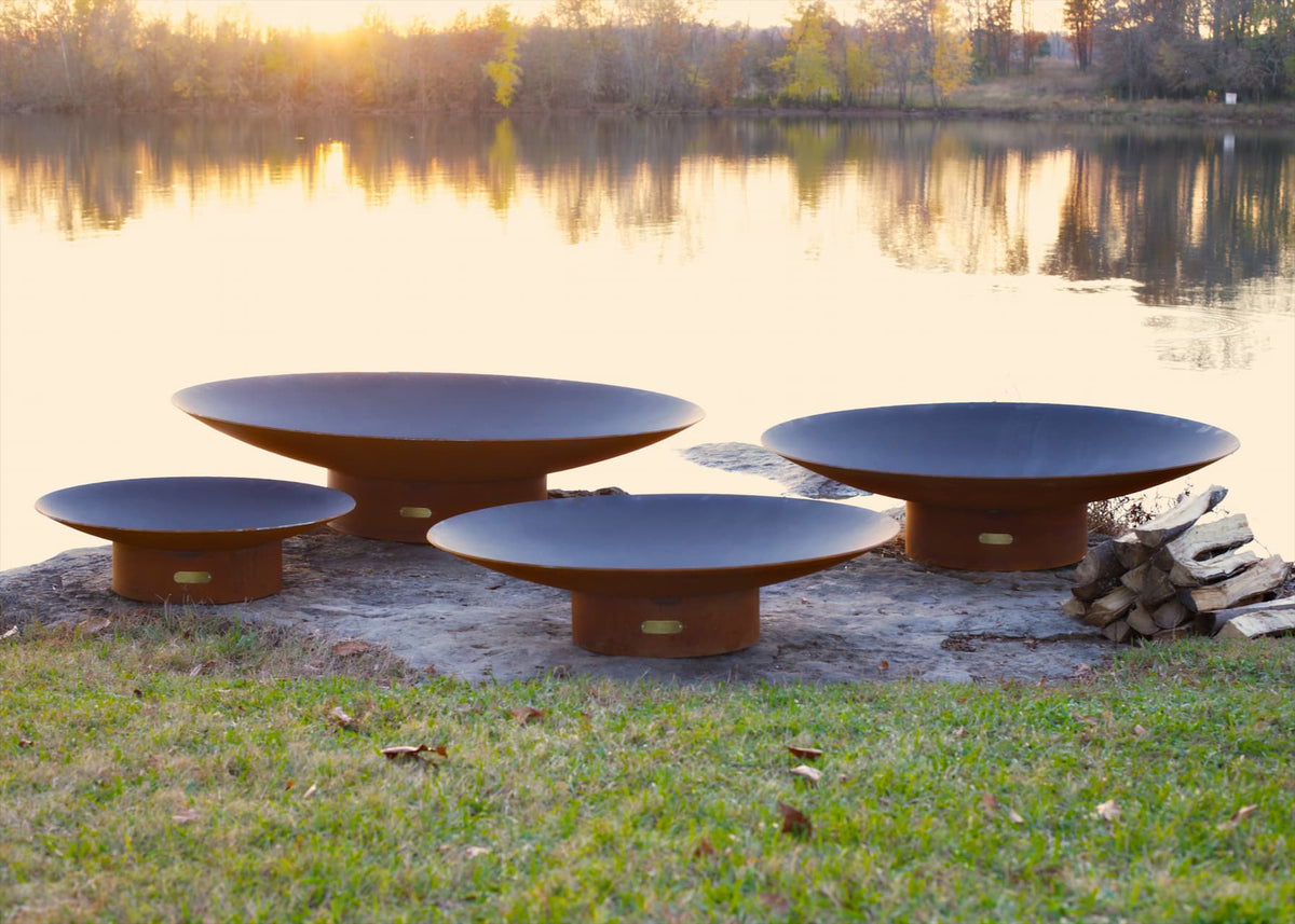 Side profile of all four sizes of the Asia Steel Fire Pit on rocky shore near dock.