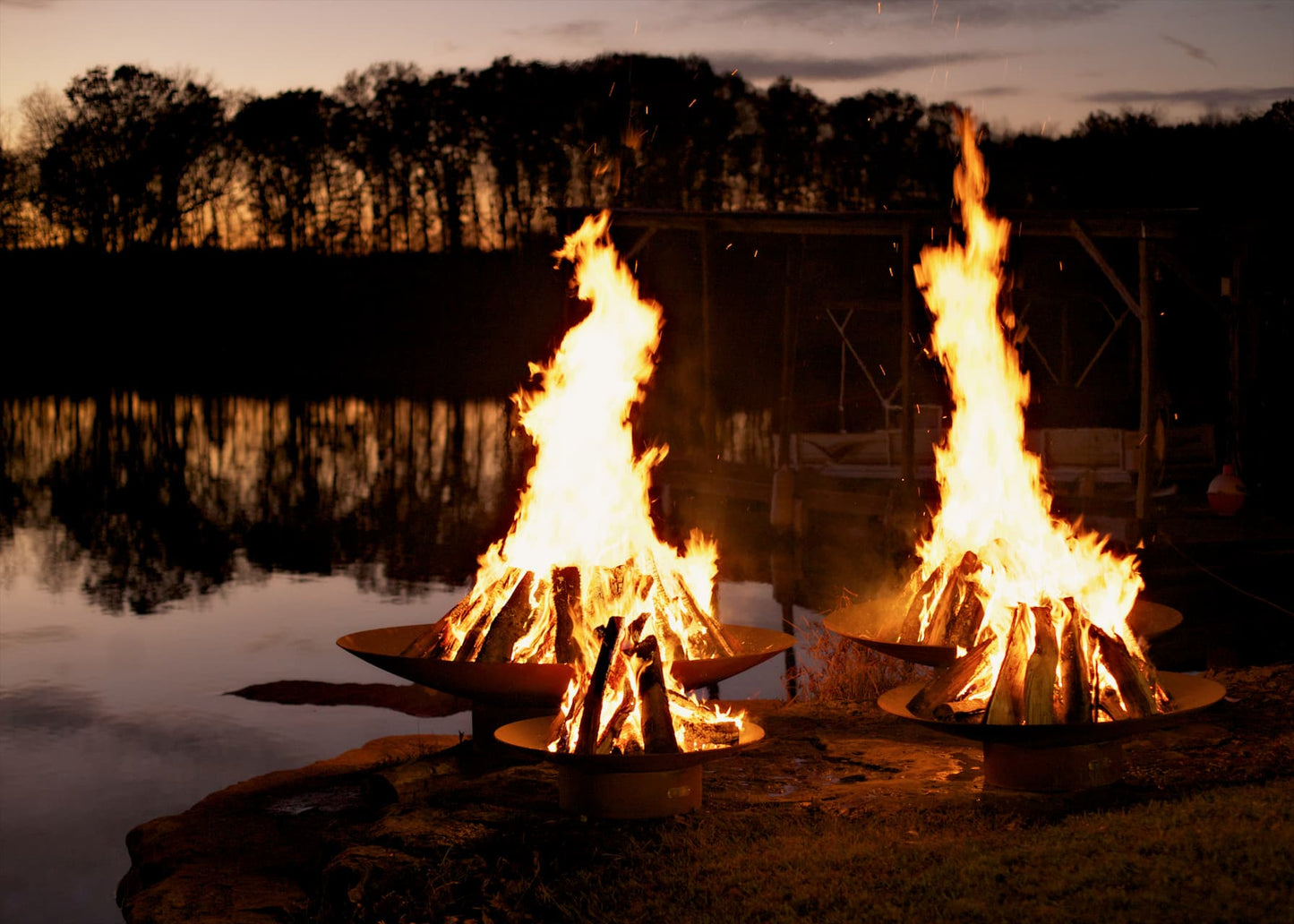 Multiple sizes of the Asia Carbon Steel Fire Pit glowing in the night near water's edge. All made in the USA by Fire Pit Art.