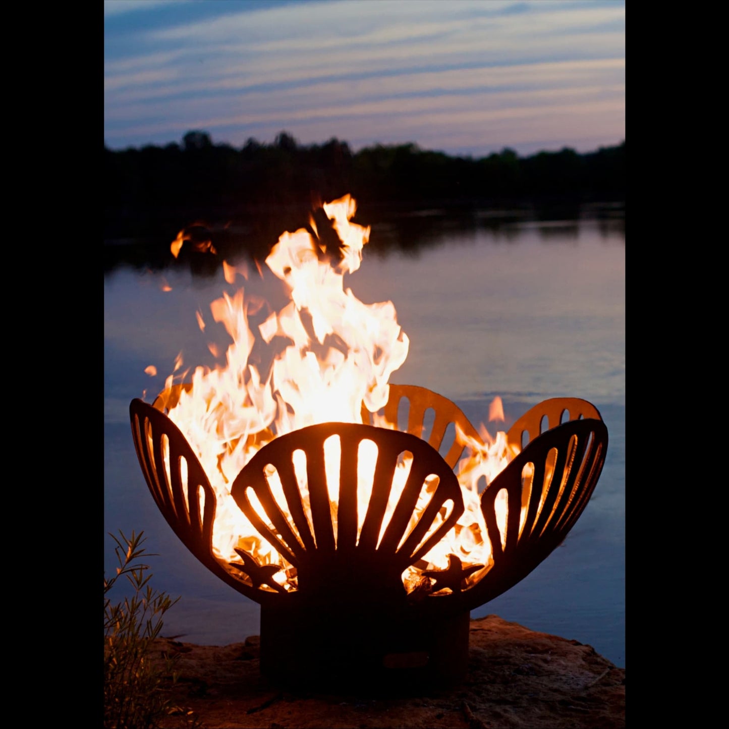 Barefoot Beach 42" Carbon Steel Fire Pit with flames burning brightly against a lake and sunset sky.
