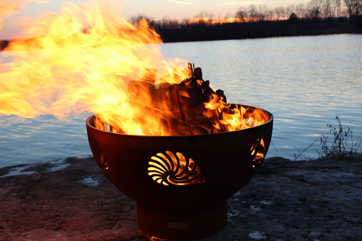 Beachcomber 36" steel fire pit lit near calm lake with flame illuminating shell cutouts. Made in the USA by Fire Pit Art.