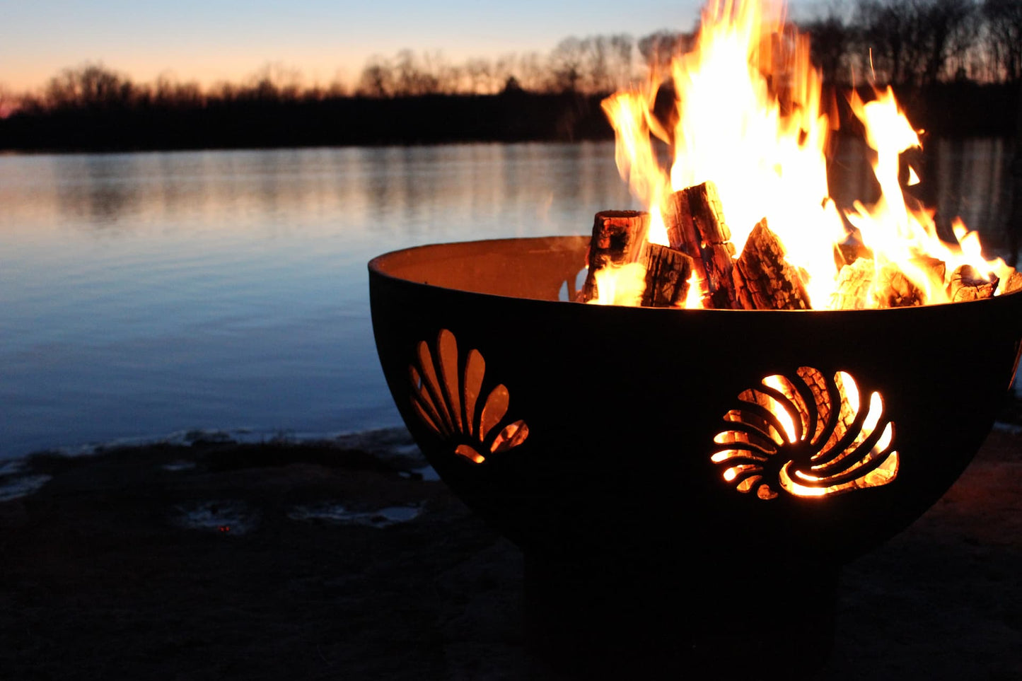 Glowing Beachcomber 36" steel fire pit with fire against a lake backdrop as the sun sets.