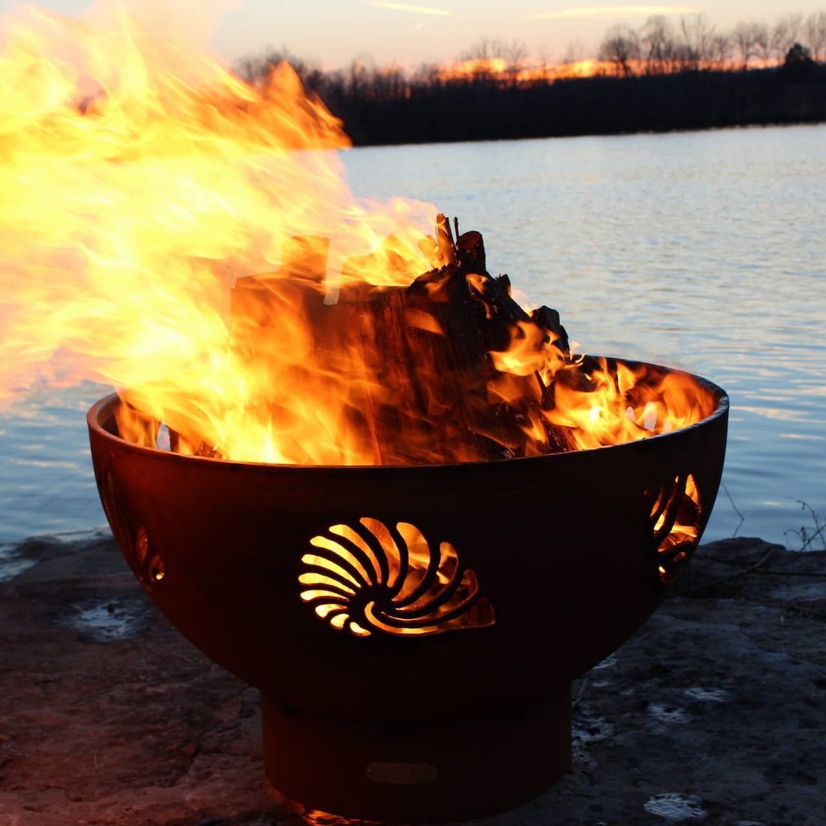 Beachcomber 36-inch carbon steel fire pit with seashell cutouts lit by fire at sunset. Made in USA by Fire Pit Art.
