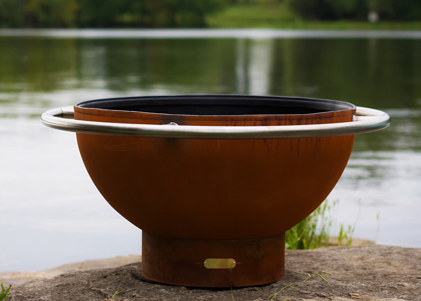 Close-up of stainless steel ring on Bella Luna 48" carbon steel fire pit with lake in the background. Made in USA by Fire Pit Art.
