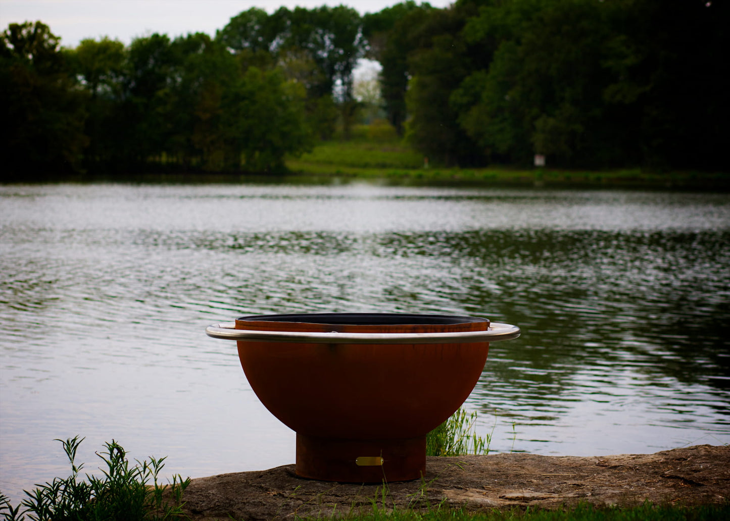 Bella Luna 48" carbon steel fire pit in a dusk setting by calm lake water. Made in USA.