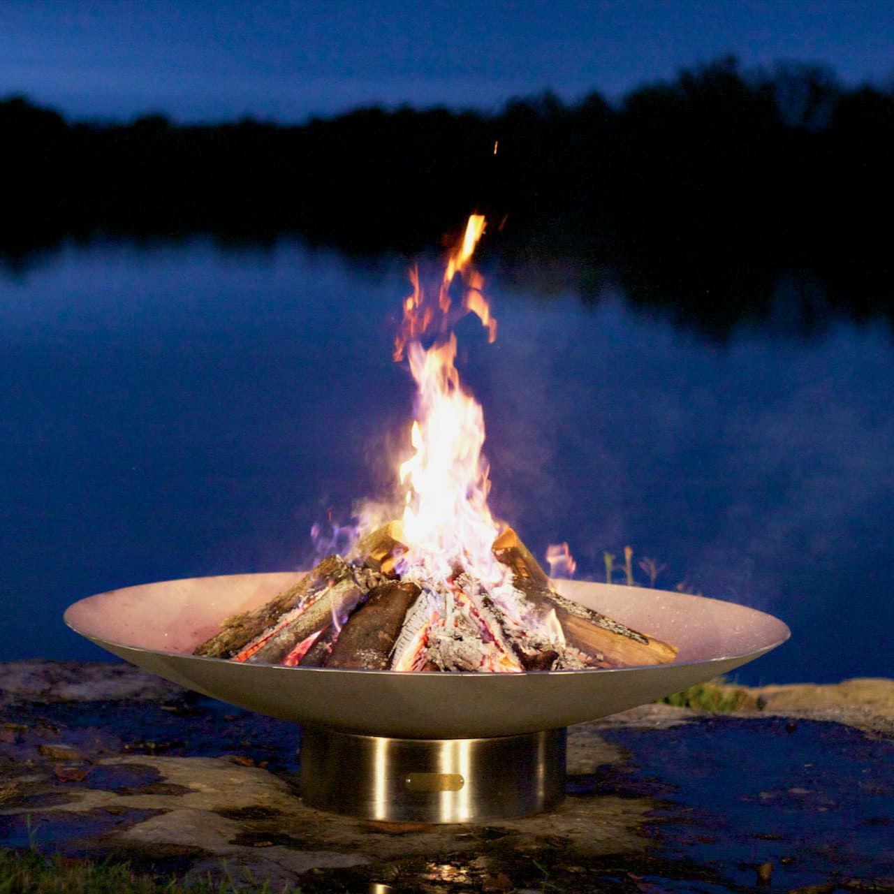 Bella Vita stainless steel fire pit with flames burning brightly in a metallic bowl by a body of water at night. Made by Fire Pit Art in the USA.