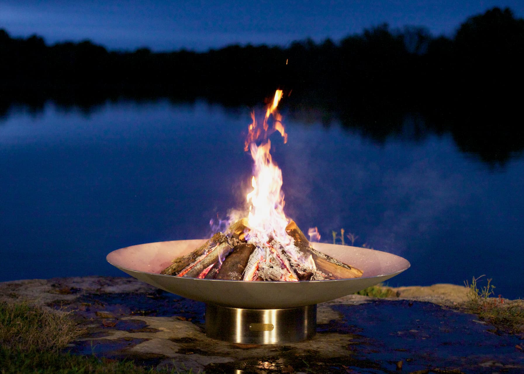 Fire Pit Art's Bella Vita stainless steel fire pit in evening with tall flames and lake backdrop.