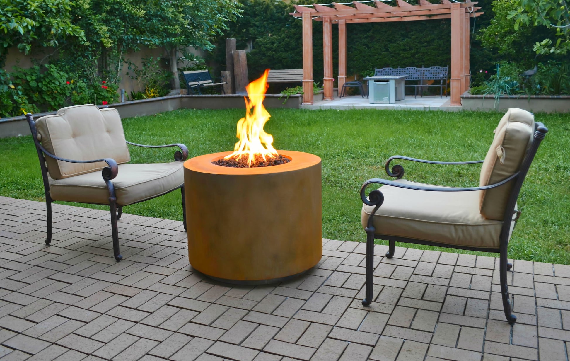 Beverly Corten Steel Propane Fire Pit lit in a backyard with patio chairs and green lawn.