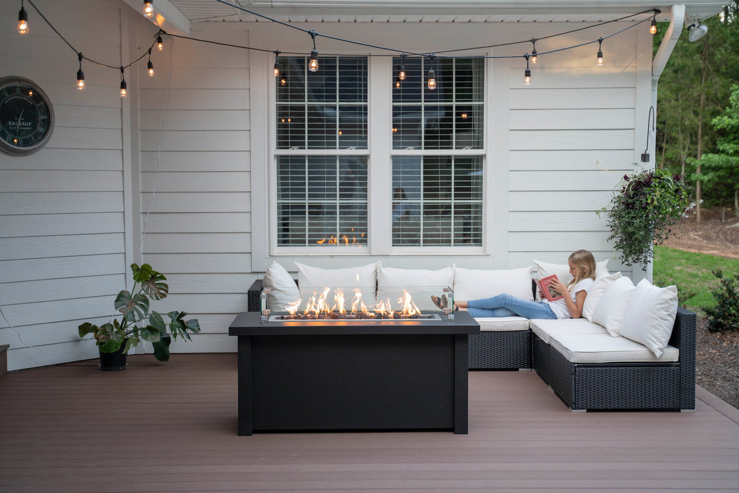 Front view of lit Carolina Evening Companion Propane Fire Table - Linear in Graphite at dusk on patio with girl reading in background (wind guard sold separately).