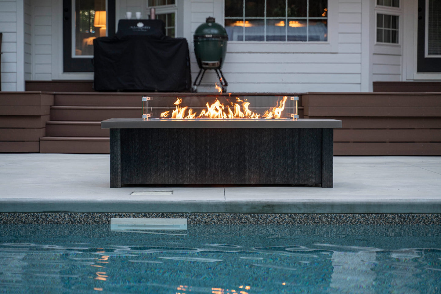 Front view of lit Carolina Evening Companion Propane Fire Table - Linear in Brushed Black with pool in foreground (wind guard sold separately).