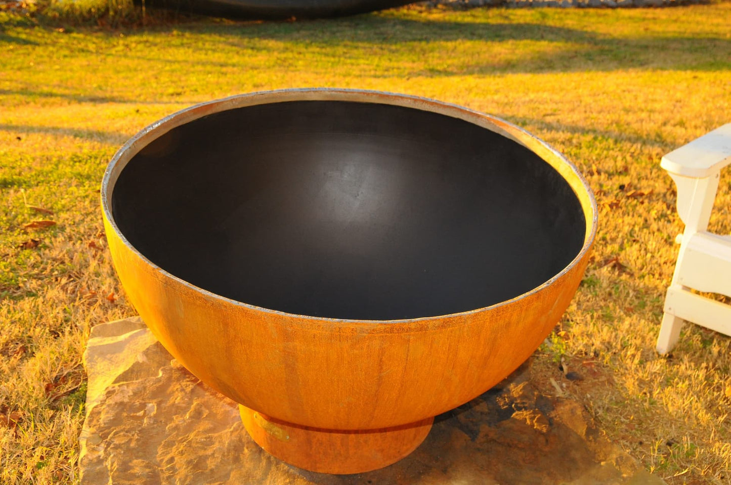 Fire Pit Art's Crater 36" carbon steel fire pit pictured at an angle with open interior and Adirondack chair in background. Made in USA.