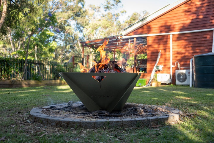 The Delta black steel fire pit lit in a backyard patio ring with a red house and trees in background.