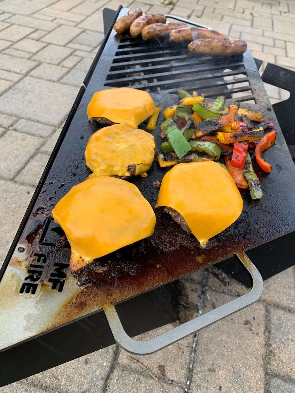 Cheeseburgers, peppers, and sausage grilling on MF Fire Delta fire pit cooktop with open flame underneath.