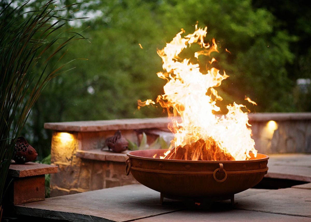 Emperor fire pit near a lakeside patio with warm firelight and greenery in background. Made in USA by Fire Pit Art.
