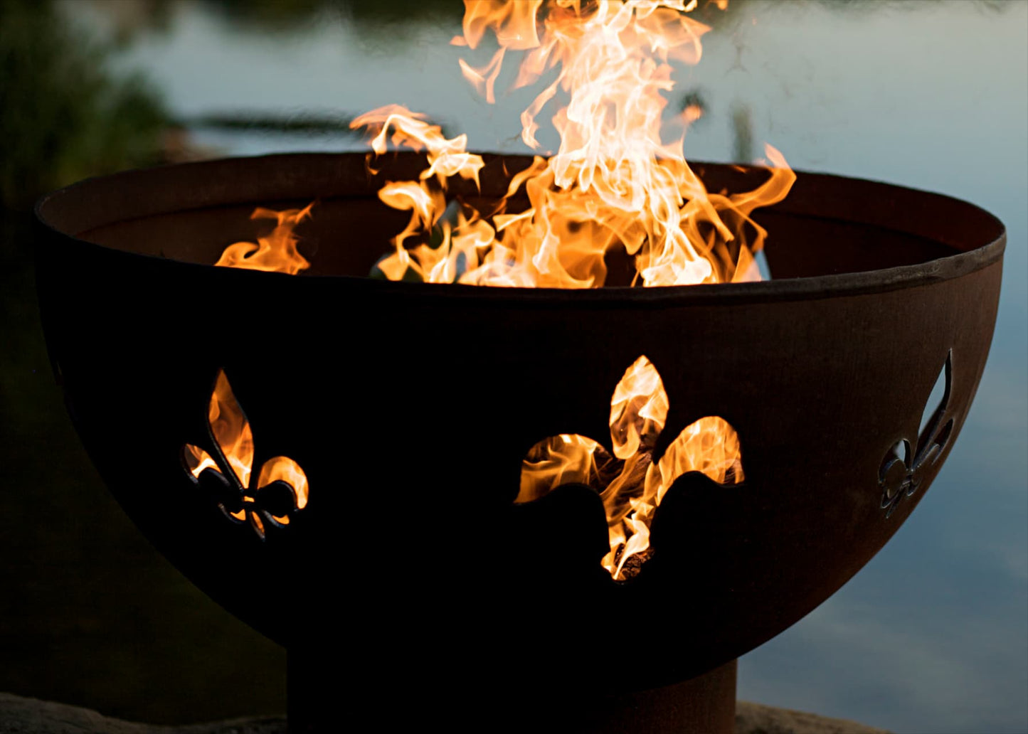Fire Pit Art's Fleur de Lis 36" carbon steel fire pit glowing brightly by the water’s edge with visible flame and elegant cutouts.