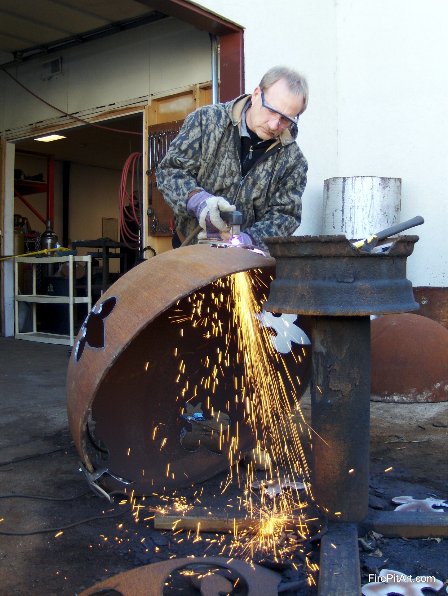 Fire Pit Art Owner and Artisan Rick Wittrig welding Fleur de Lis carbon fire pit by hand, with sparks flying and cutout shapes visible on steel shell. Handcrafted in Tennessee, USA.