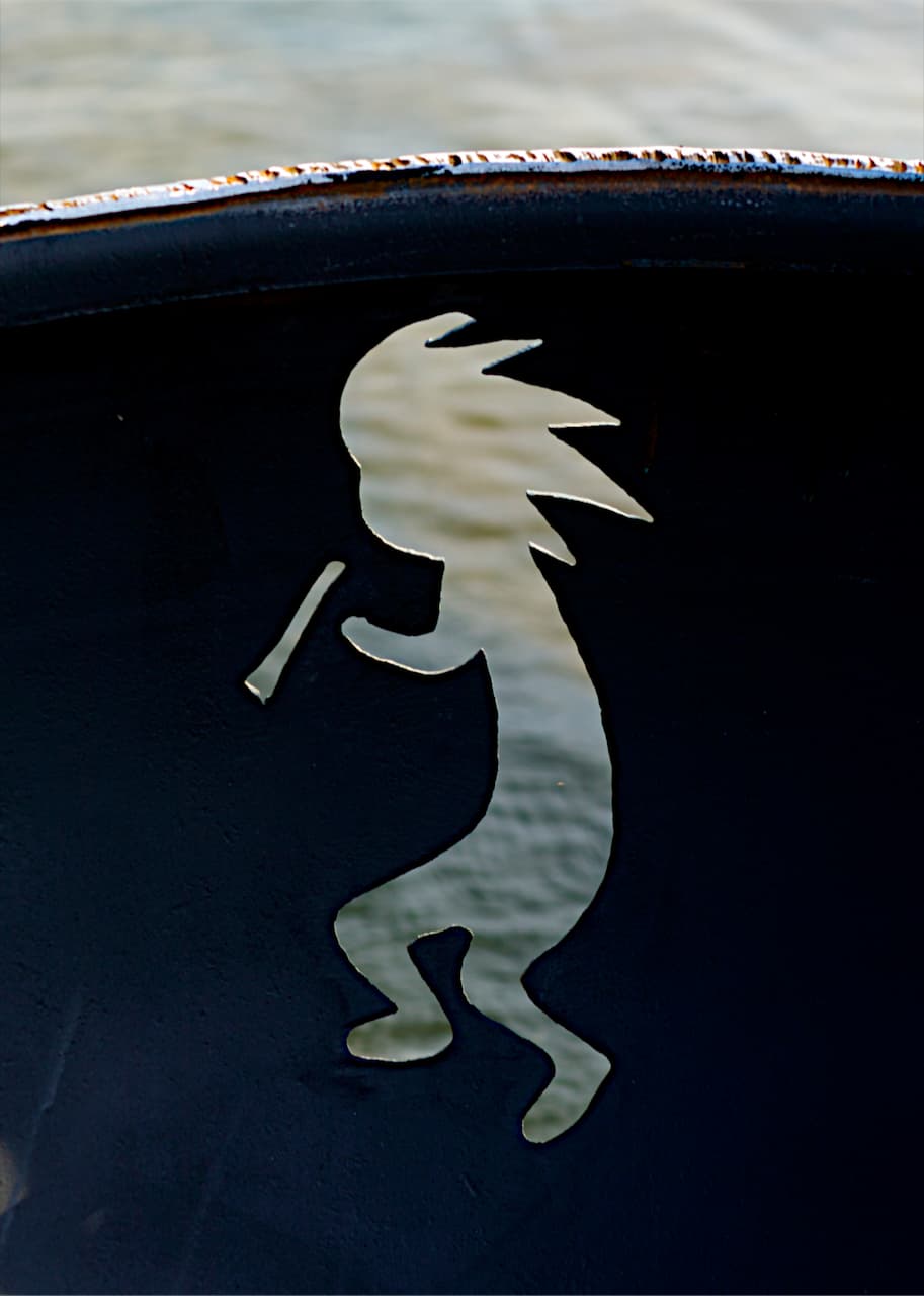 Flute-playing Kokopelli cutout on steel fire pit against water backdrop.
