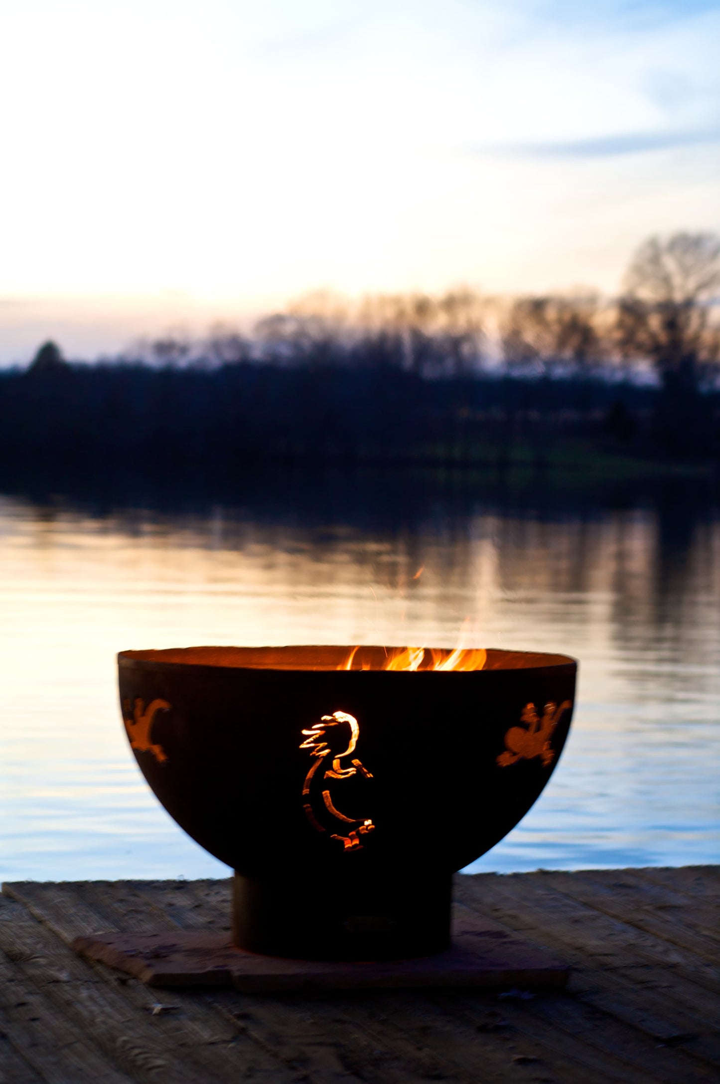 Kokopelli 36" carbon steel fire pit glowing beside a calm lake at dusk. Made in the USA by Fire Pit Art.