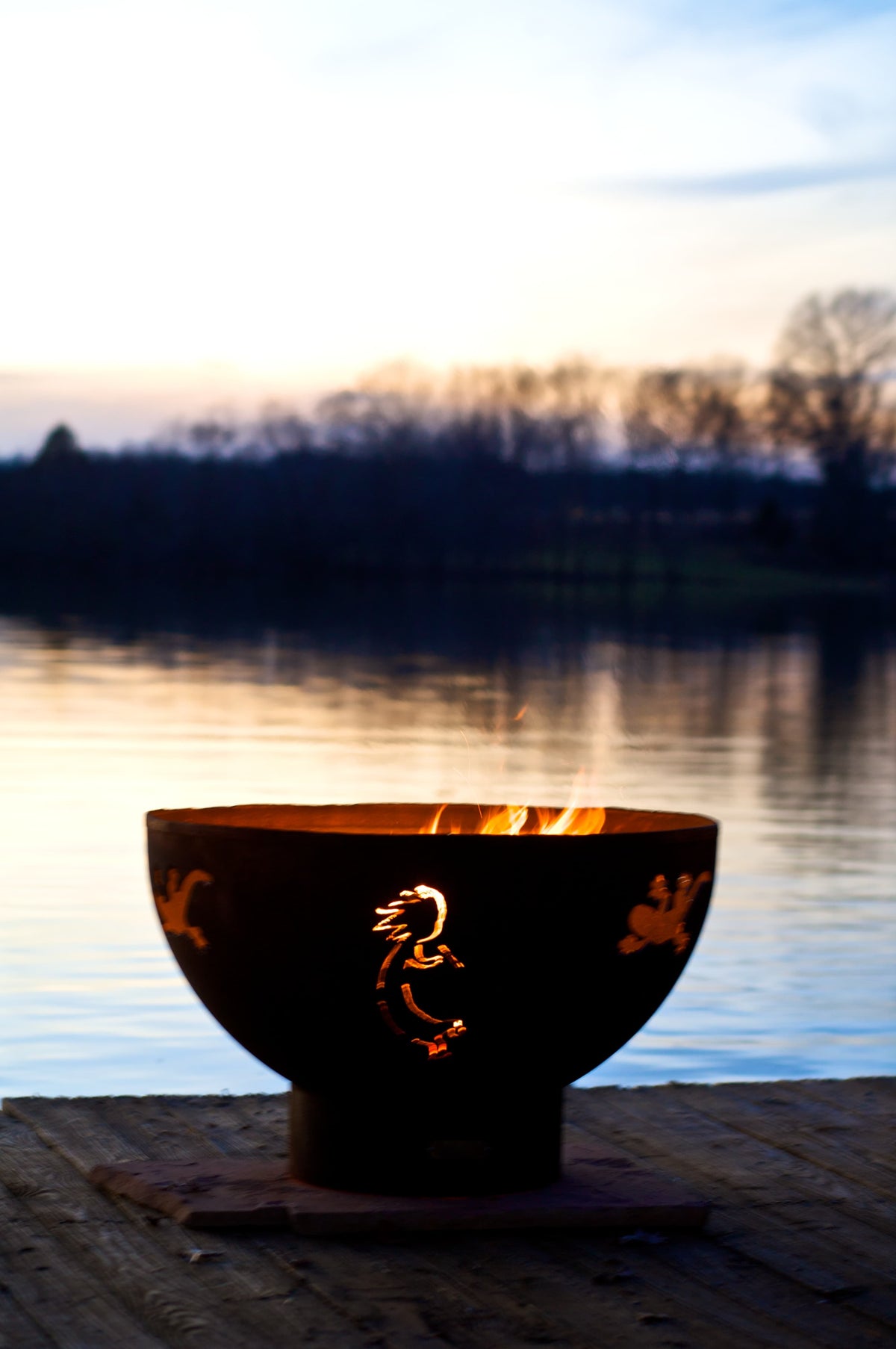 Kokopelli 36" carbon steel fire pit glowing beside a calm lake at dusk. Made in the USA by Fire Pit Art.