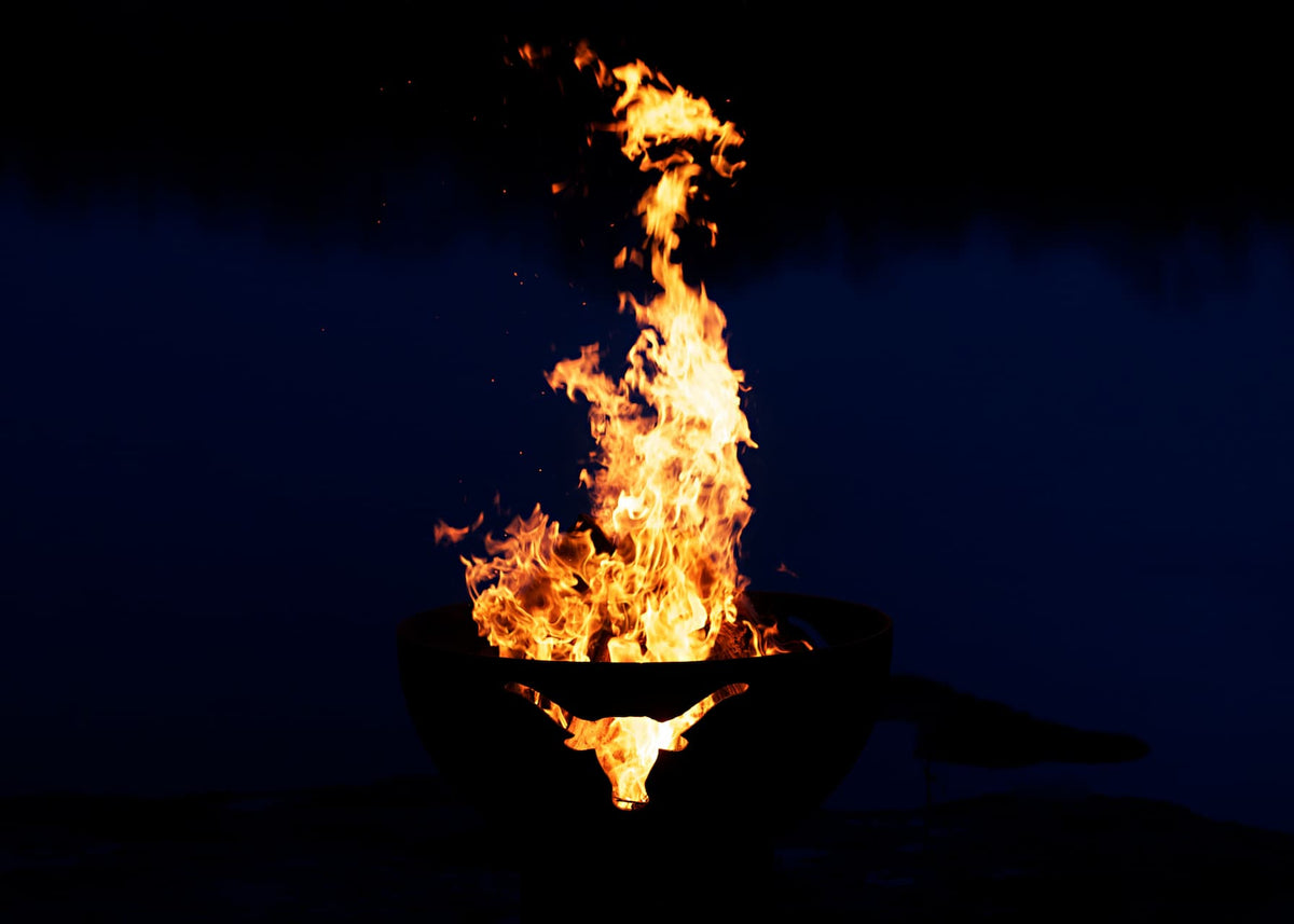 Long Horn 36" carbon fire pit blazing by water with longhorn cutout glowing on a very dark night.