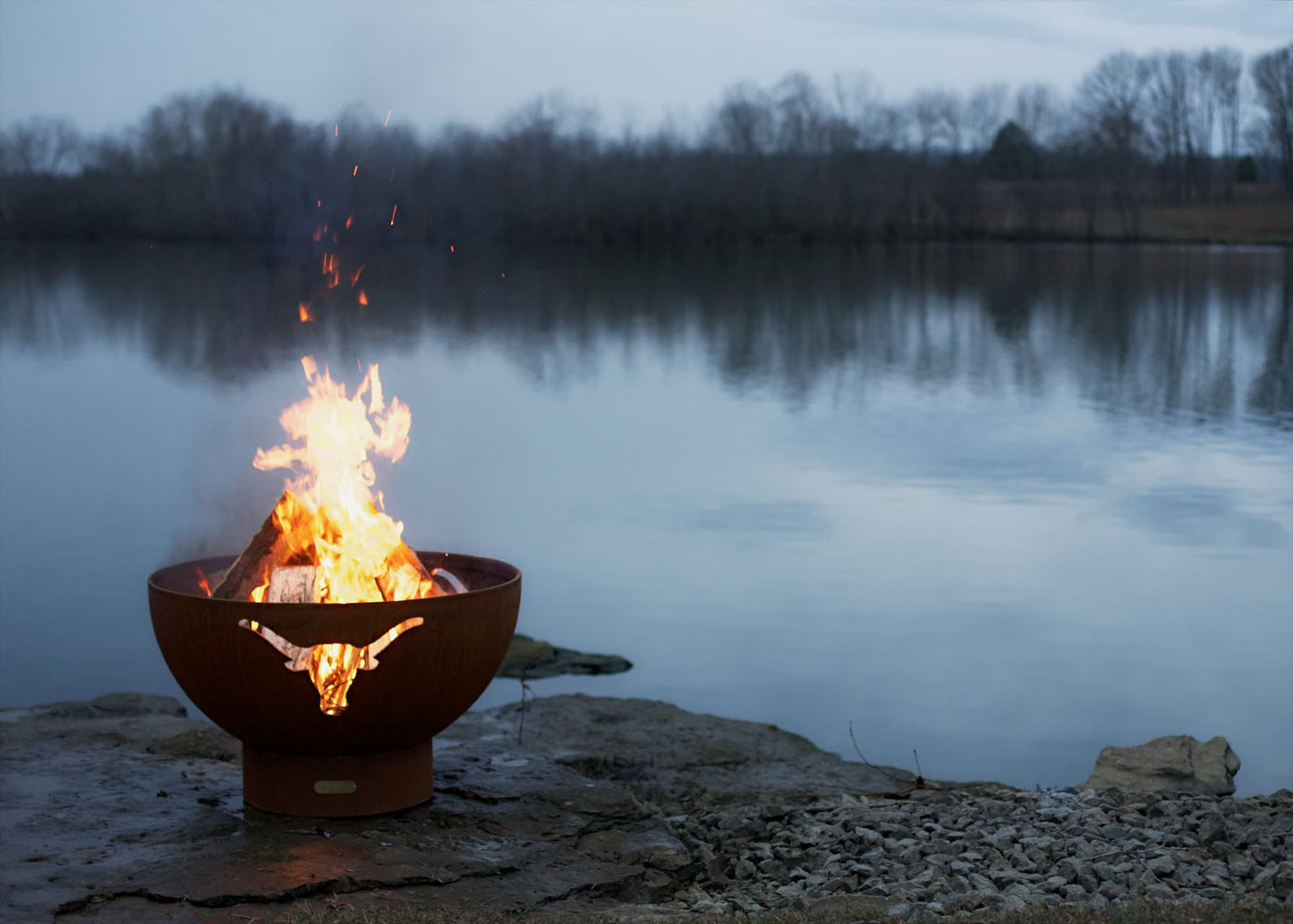 Long Horn 36" carbon steel fire pit with flames by a body of water at dusk.