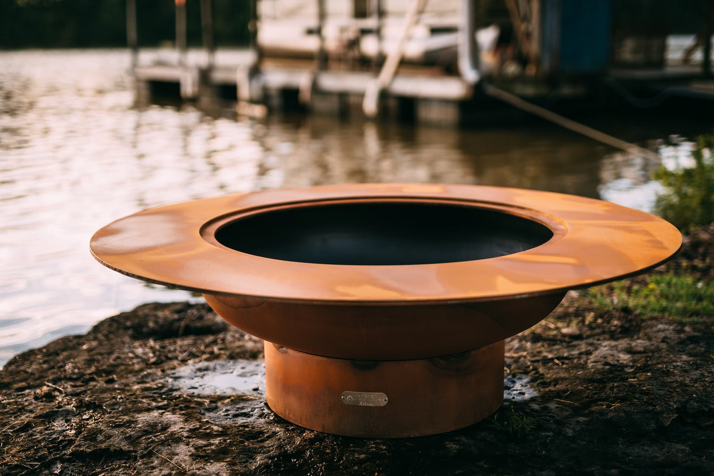 Magnum 54" carbon steel fire pit on shoreline near a dock. Made in USA by Fire Pit Art.