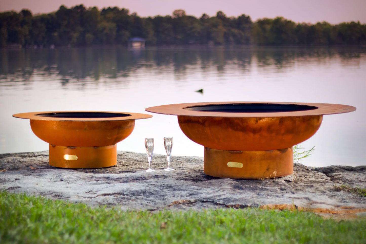Saturn 41" steel fire pit on left next to Magnum 54" steel fire pit on right with lake in background.