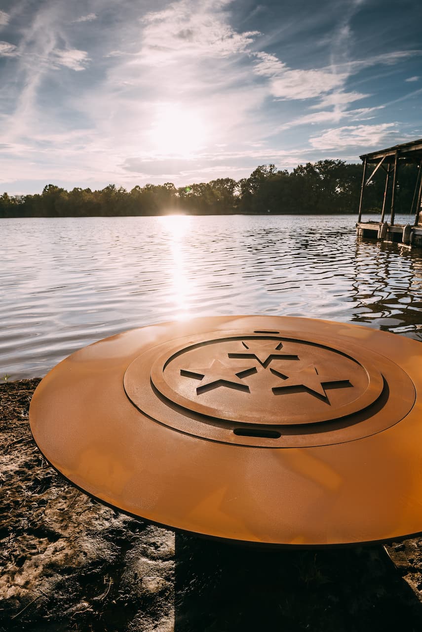 Side profile of the Magnum 54" carbon steel fire pit, with three star lid on top, next to a sunny lake.