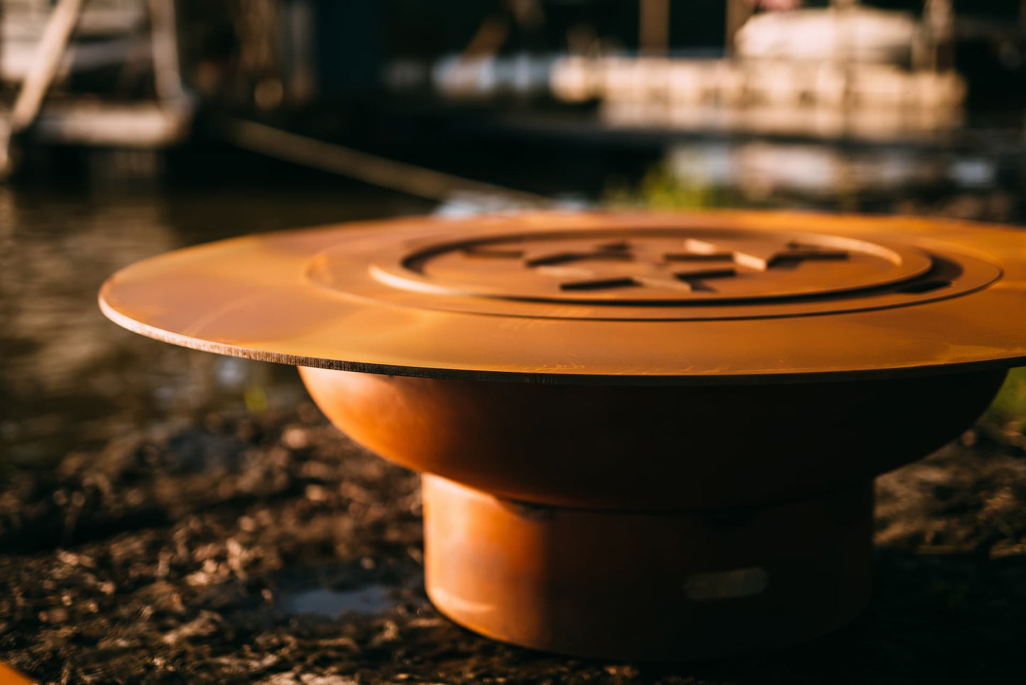 Closeup of the Magnum 54" carbon steel fire pit with beautiful three-star lid on top of it. A lakeside dock is blurred in the background.