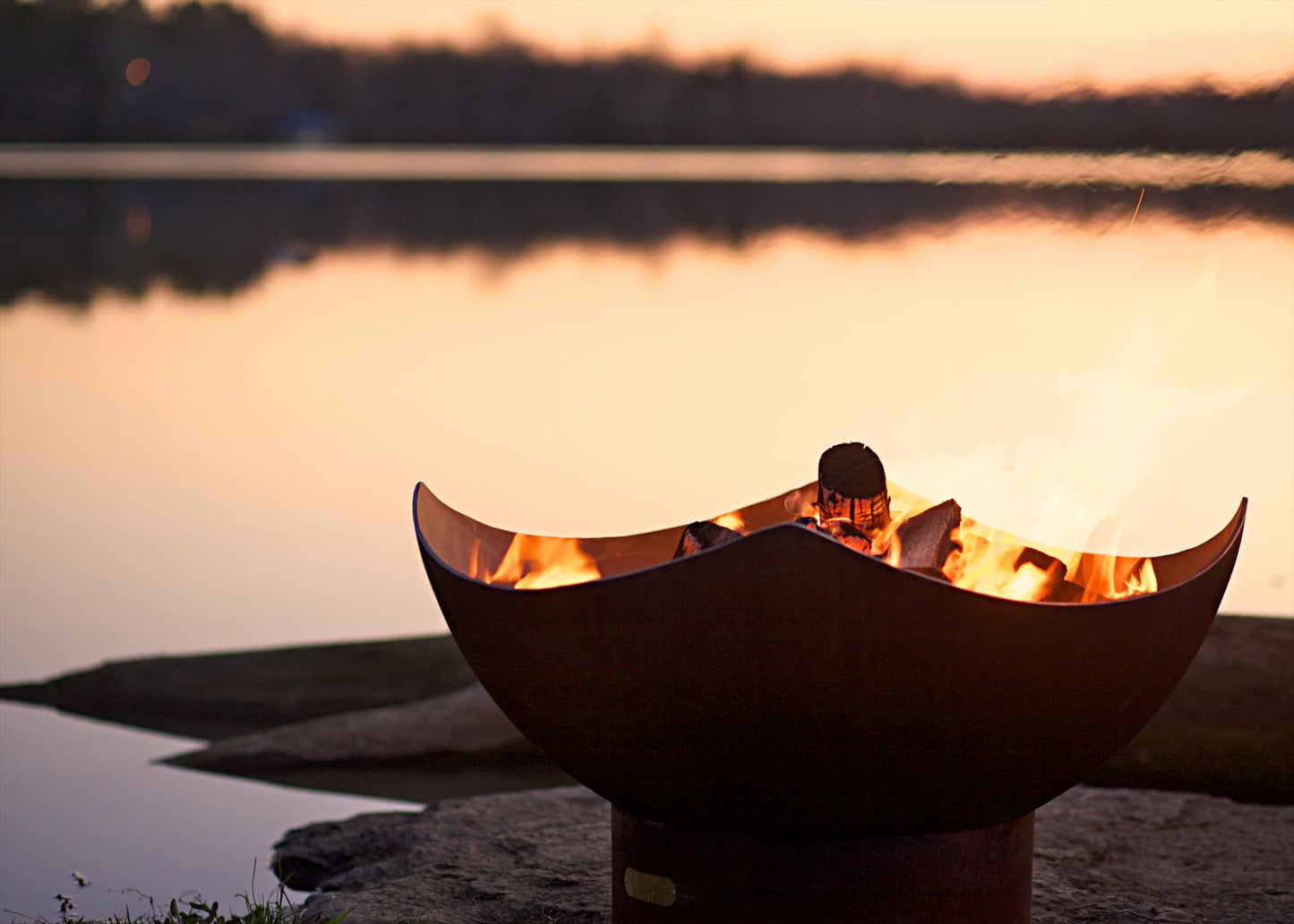 Glowing wood burning in Manta Ray 36" carbon steel fire pit on a lake at sunset.