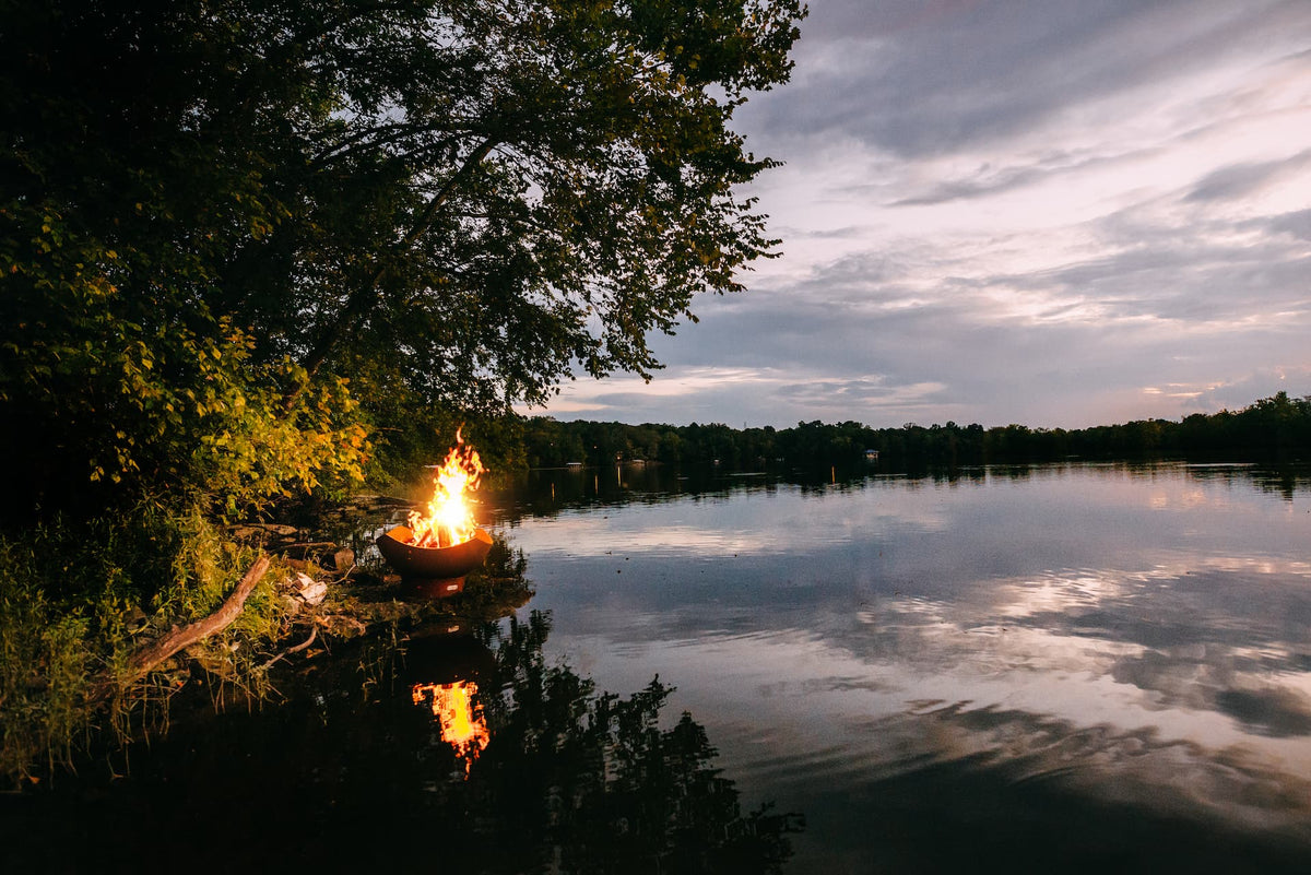 Manta Ray 36" carbon steel fire pit glowing at sunset by a calm lake on a stone platform.