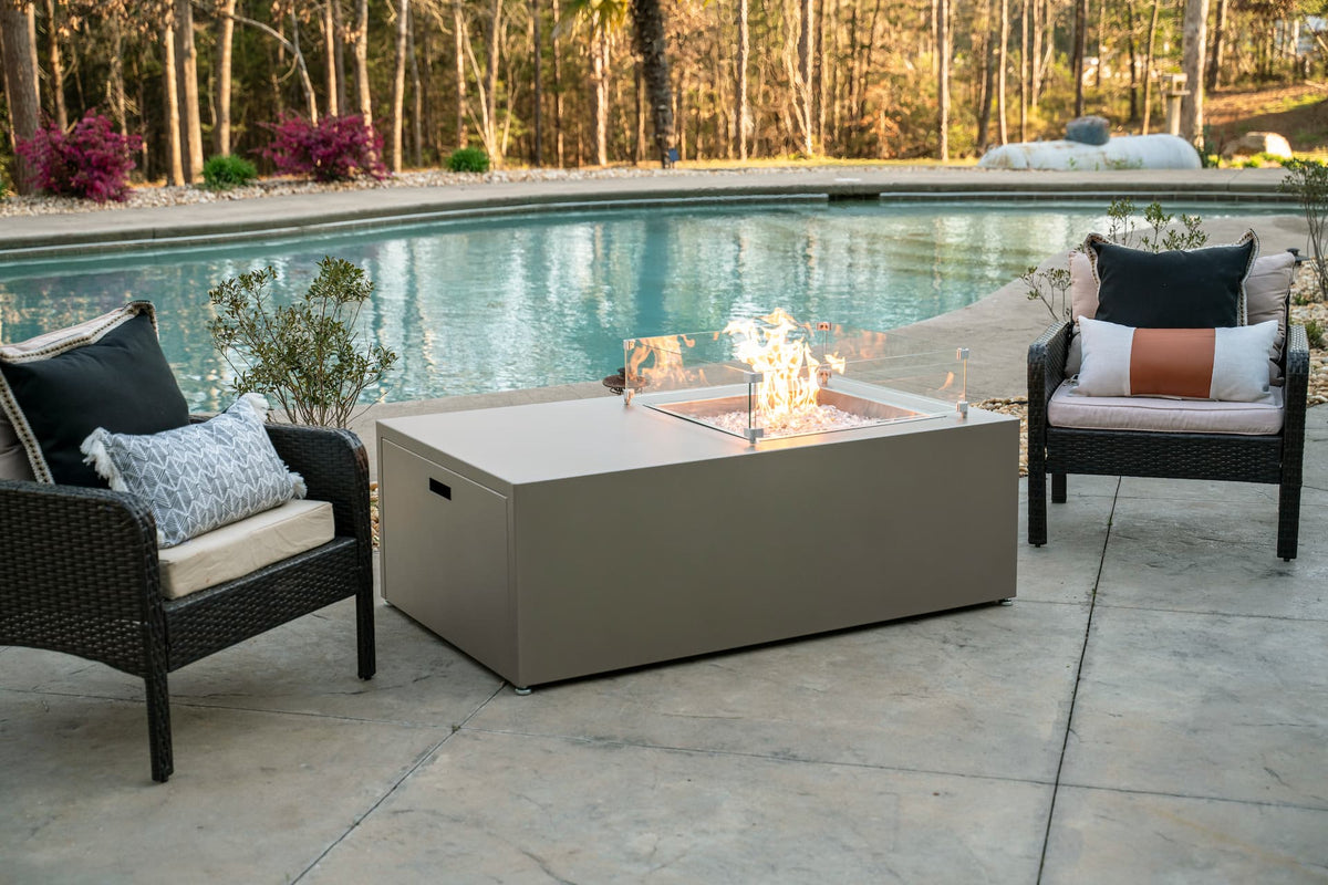 The Milan Propane Fire Table - Offset in Champagne on a patio by the pool, surrounded by wicker lounge chairs (wind shield sold separately). Shown in a retired finish β see swatches for current Endura options.