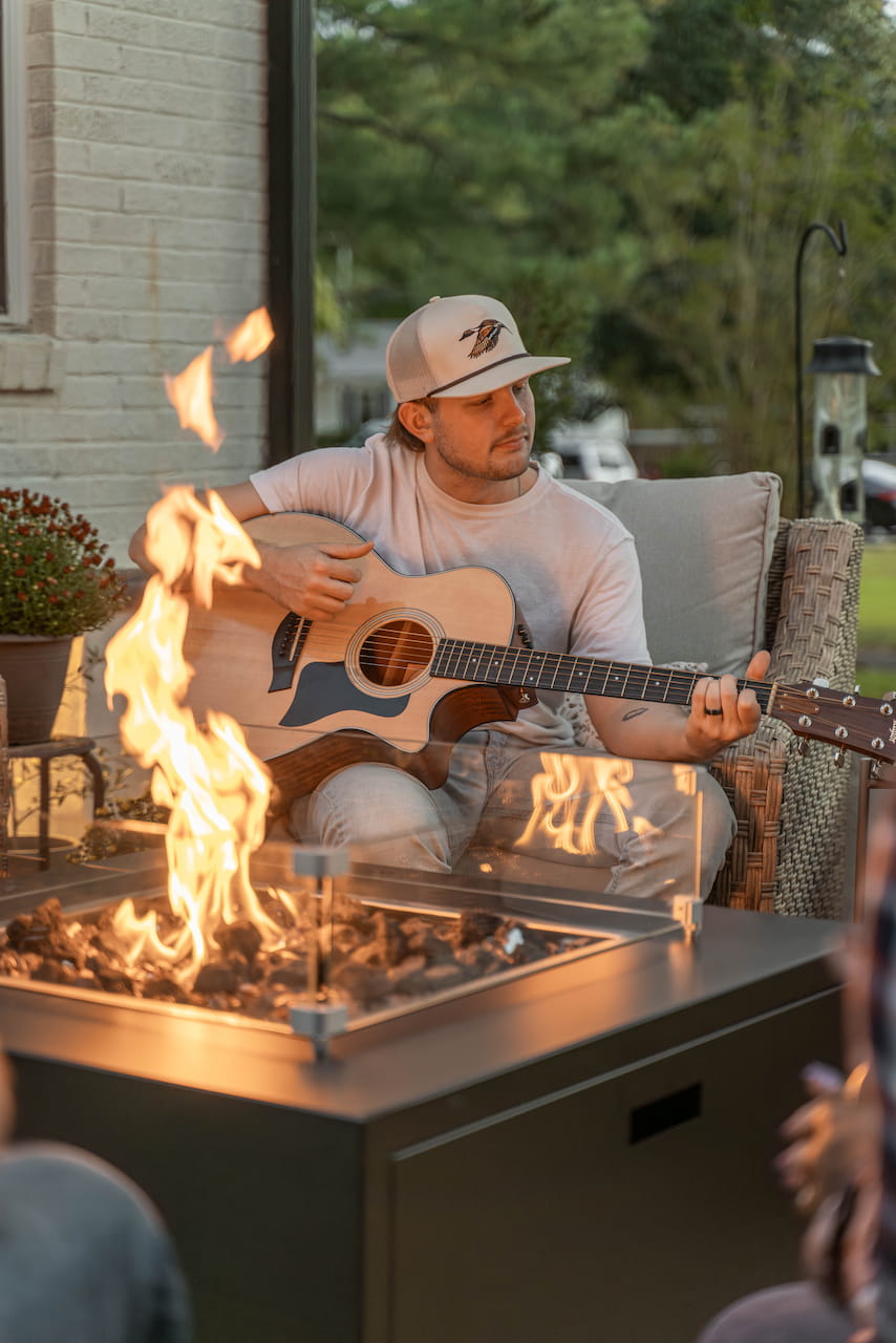 Man playing acoustic guitar next to The Milan Propane Fire Table - Square with warm flames and relaxed vibe (pictured in Endura Graphite, wind shield sold separately).