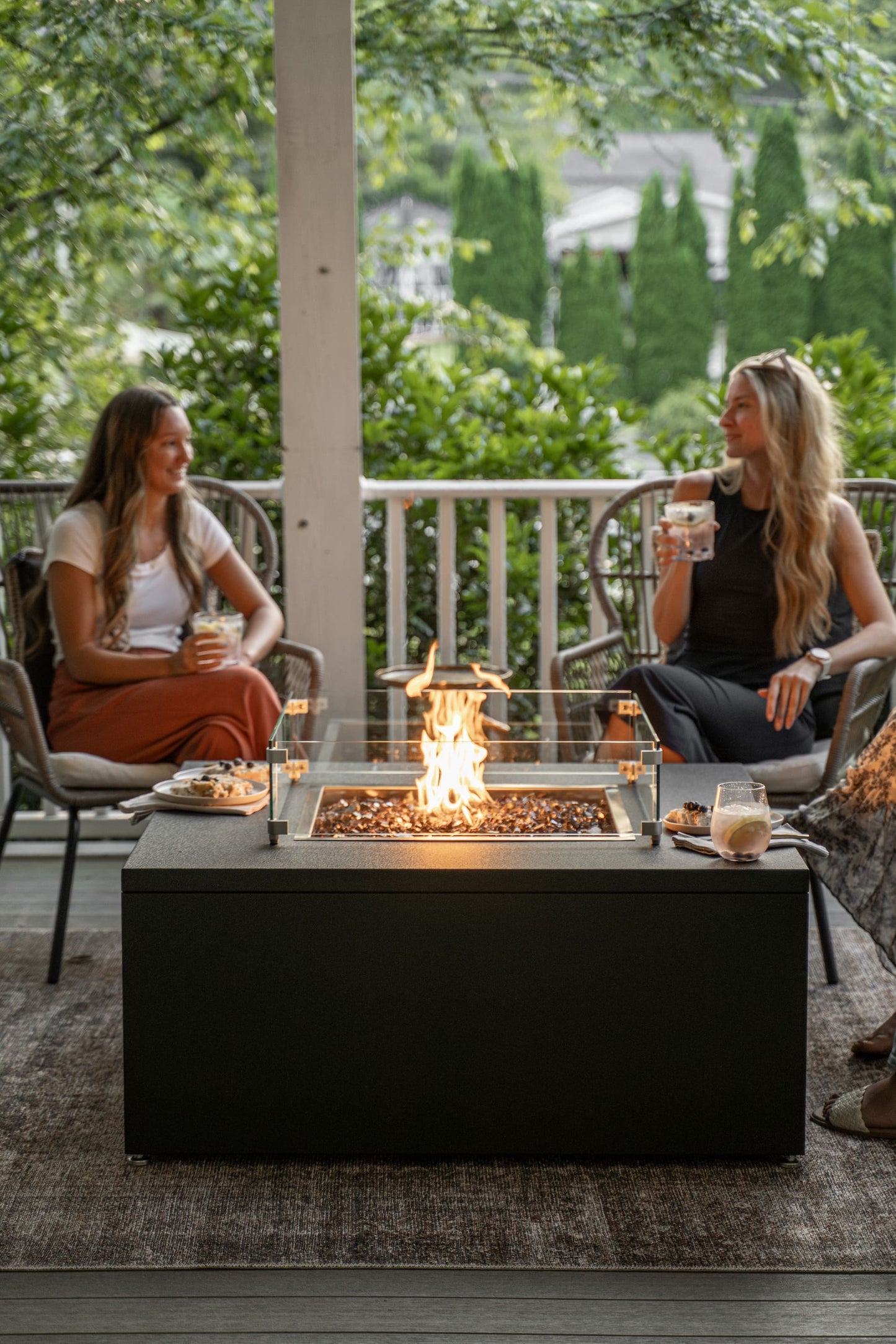 Two women sitting around The Milan Square fire pit table on a patio with greenery in the background.