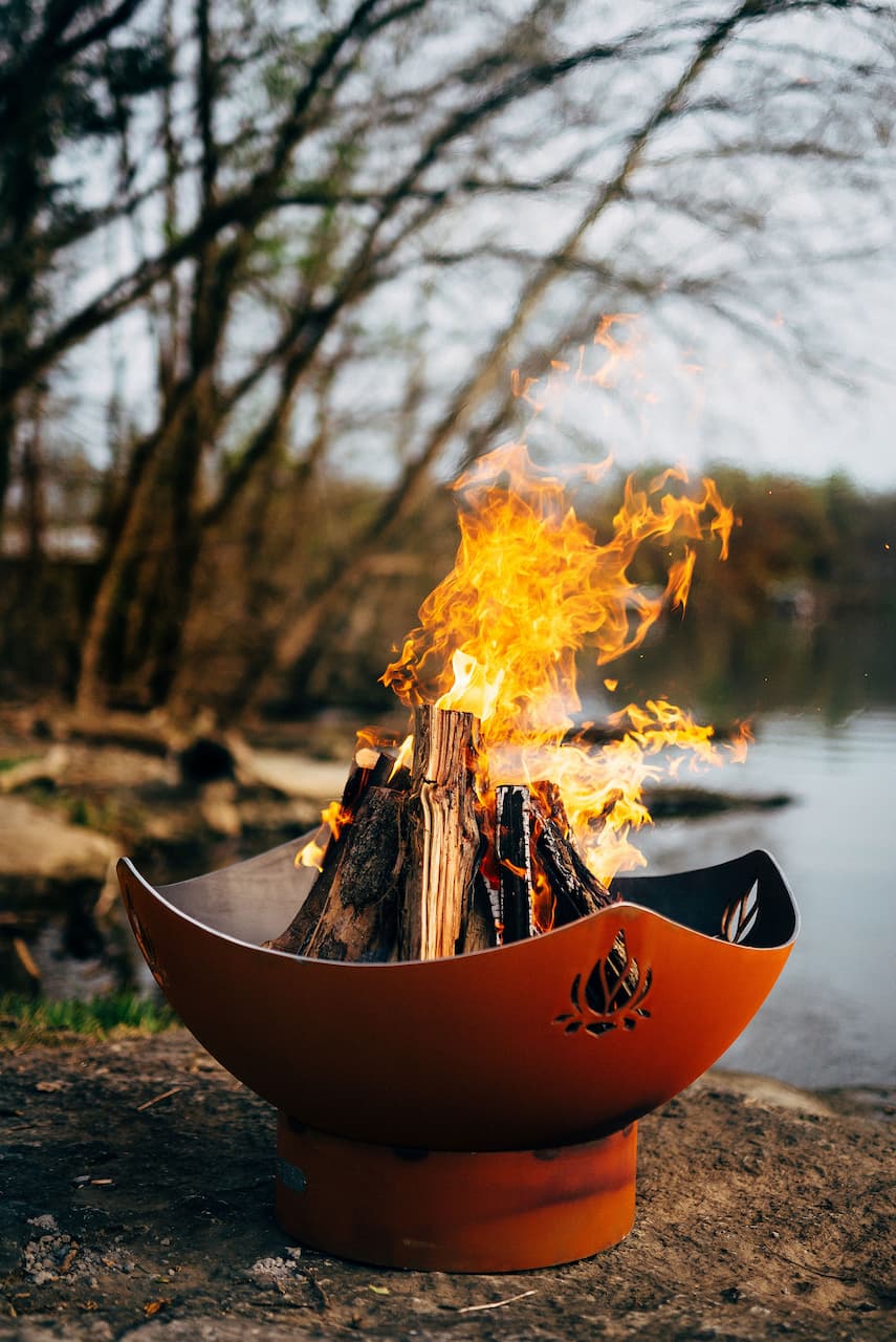 Namaste' 36" carbon steel fire pit with firewood blazing near a lake.