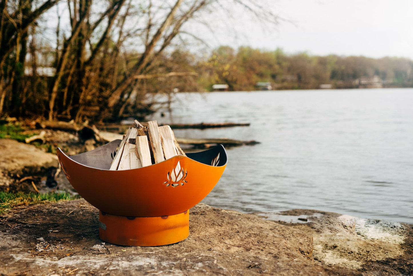 Namaste' 36" carbon steel fire pit by a lake and filled with firewood that has not yet been lit.