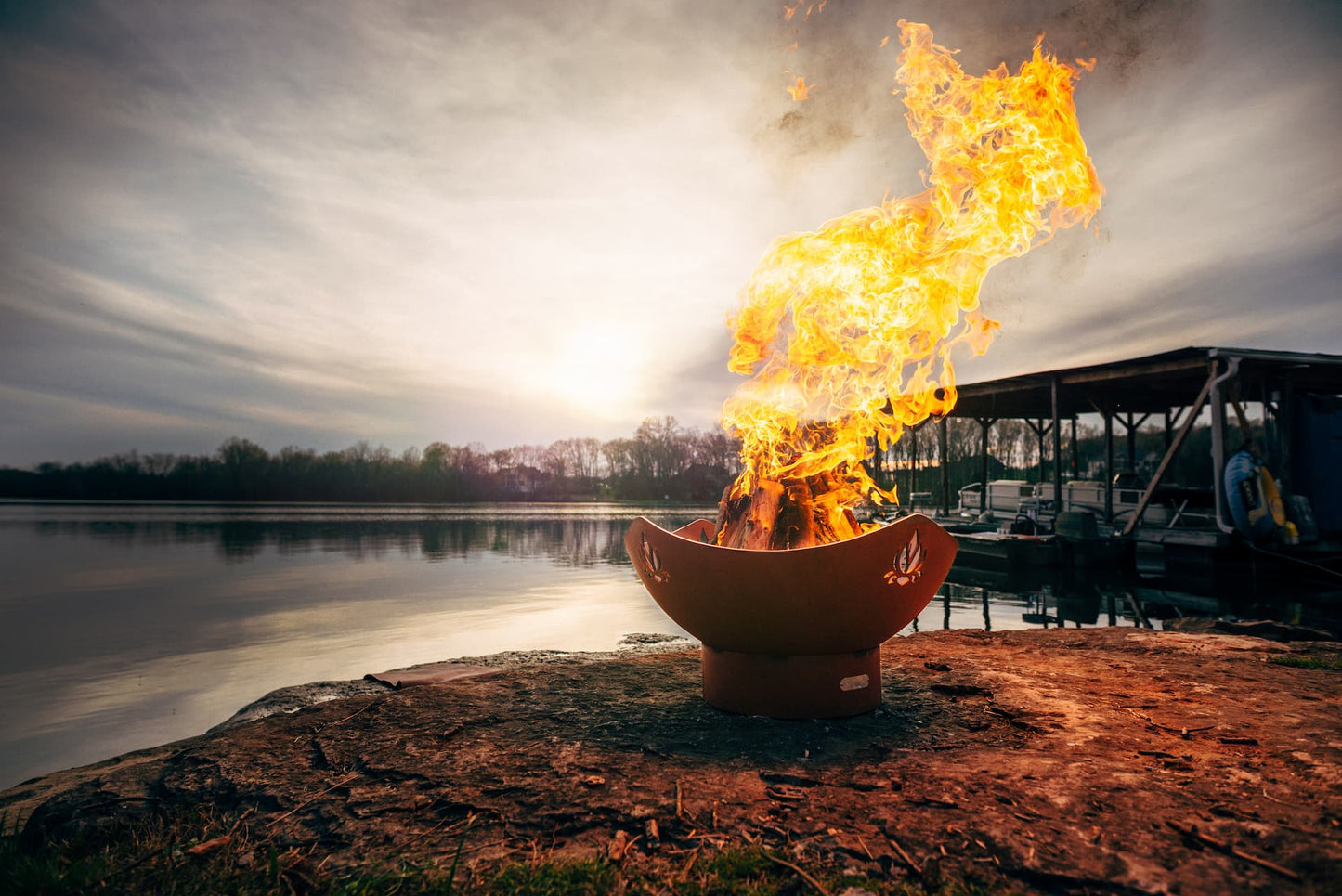 Sideview of Namaste' 36" carbon steel fire pit burning wood at dusk near a dock.