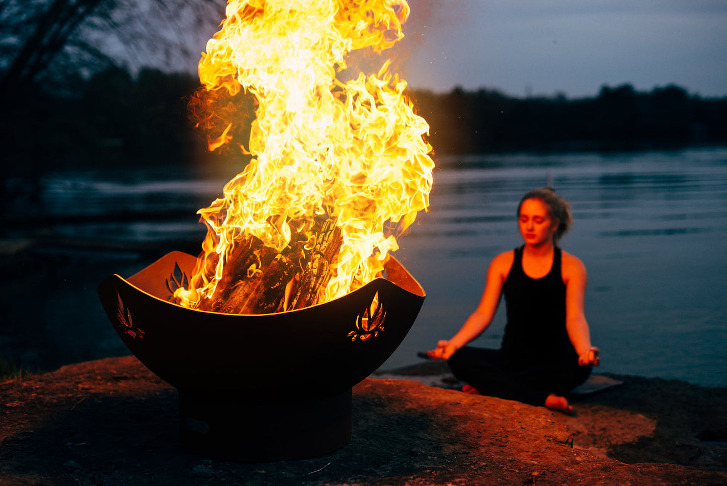 Woman meditating near Namaste' 36" carbon steel fire pit by a body of water at night.
