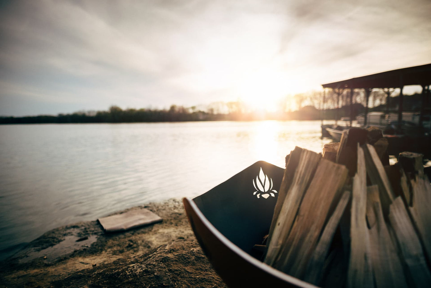 Closeup of the Namaste' 36" carbon steel fire pit with unlit wood near a lakeside dock. Made in the USA by Fire Pit Art.