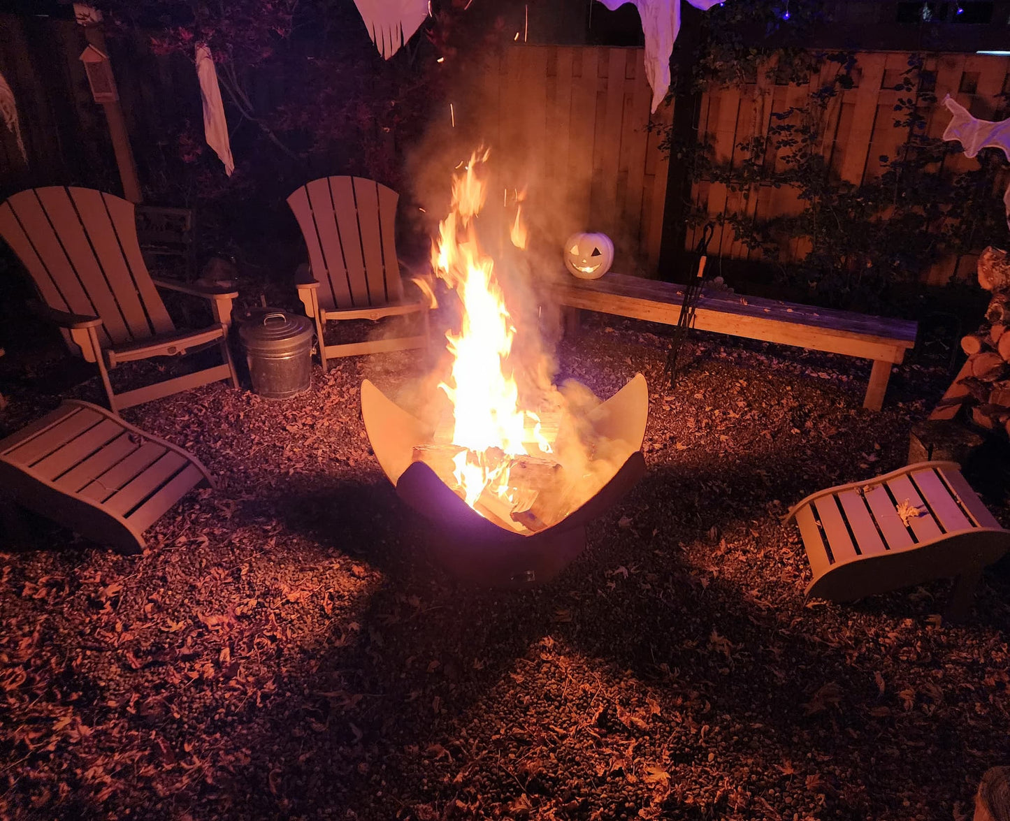 Flames rising from a blooming wood burning fire pit in a cozy backyard with Adirondack chairs and Halloween decor at night.