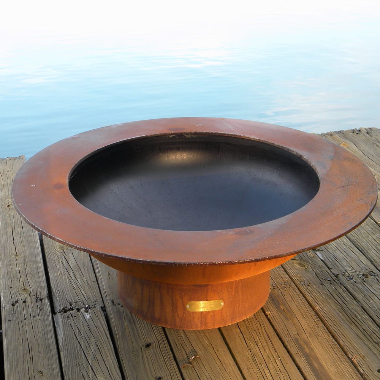 Empty Saturn 41-inch steel fire pit resting on a wooden dock, showing the smooth black interior bowl and weathered patina exterior under natural daylight.