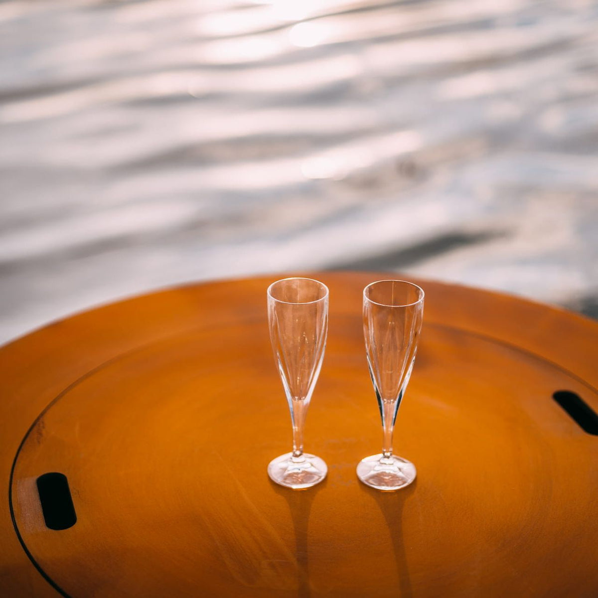 Two champagne flutes resting on the steel lid of the Saturn 41-inch fire pit, with water reflections creating a calm, elegant setting.