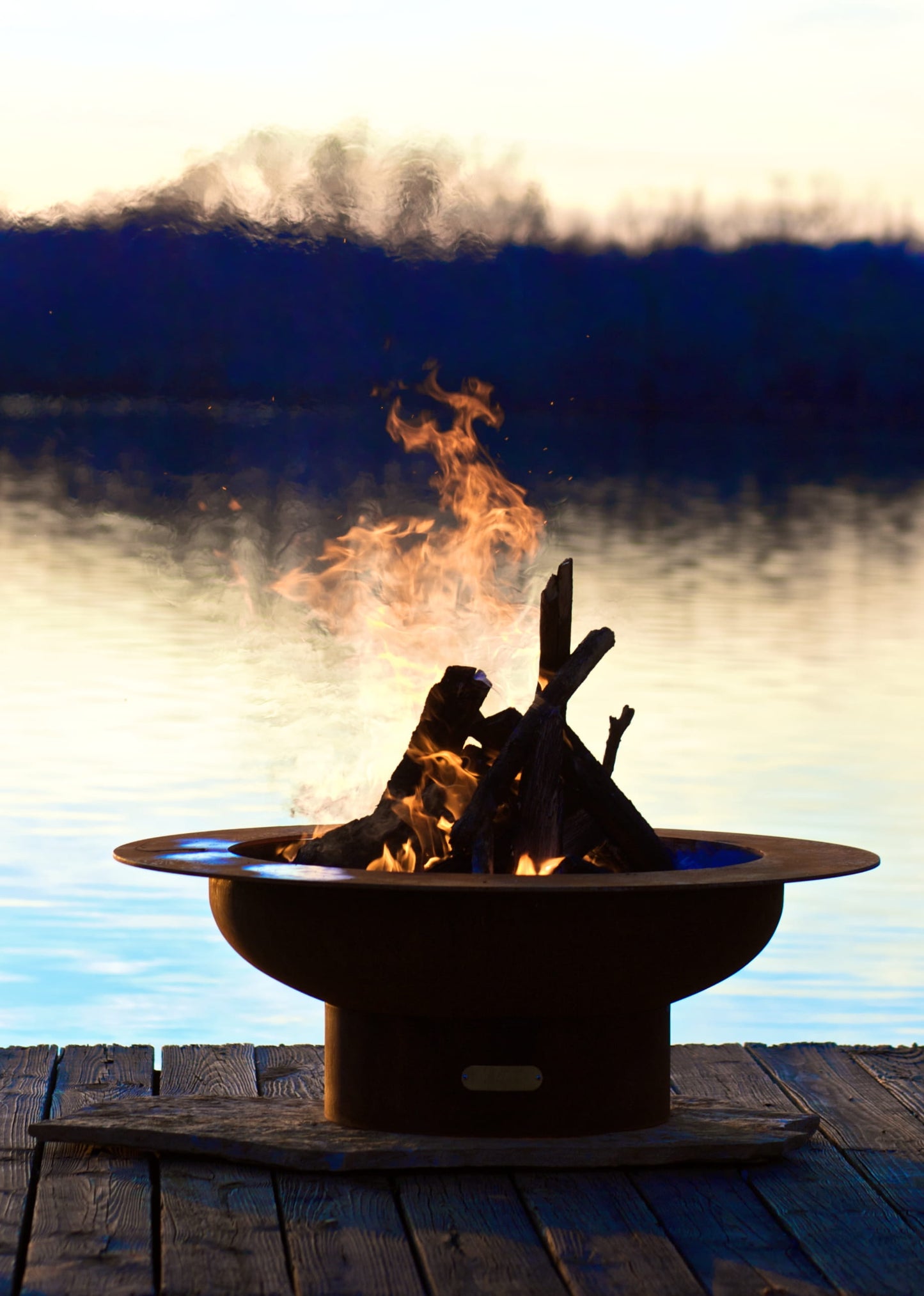 Saturn 41-inch steel fire pit lit on a dock at dusk, with flames rising above the patina steel bowl against calm lake water.