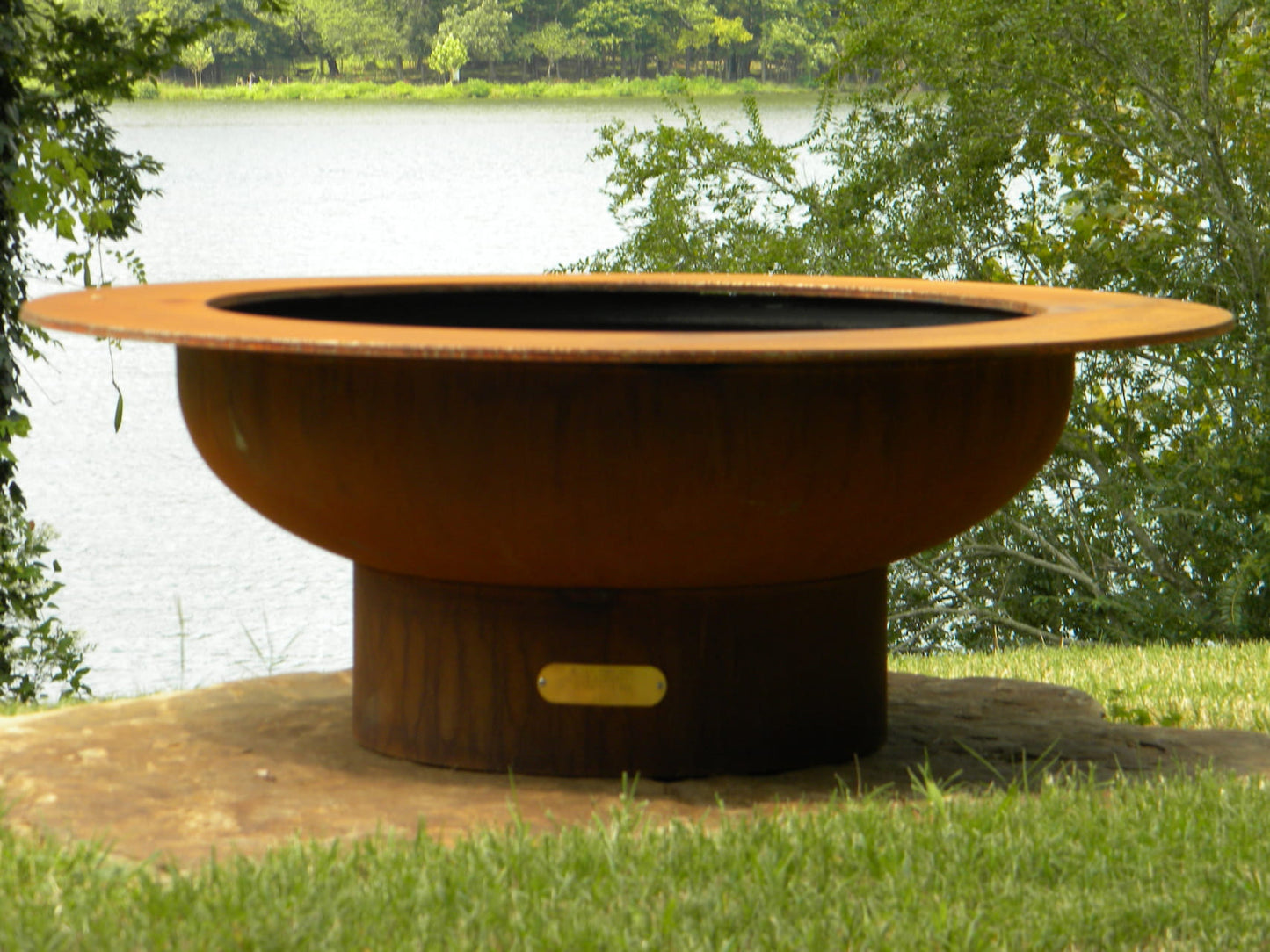 Unlit Saturn 41-inch fire pit with patina finish placed on stone slab, overlooking a peaceful lake surrounded by summer trees.