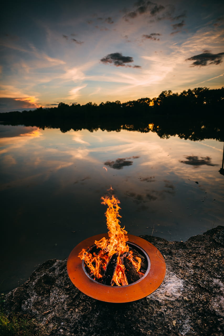 Saturn 41" carbon steel fire pit lit with tall flames, reflecting on a still lake at sunset with dramatic sky colors above the horizon. Made in the USA.