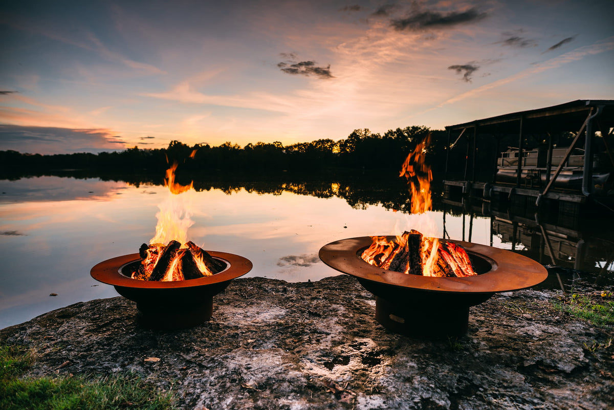 Saturn and Magnum fire pits burning side by side at sunset, showing size and flame style comparison in a lakeside setting.